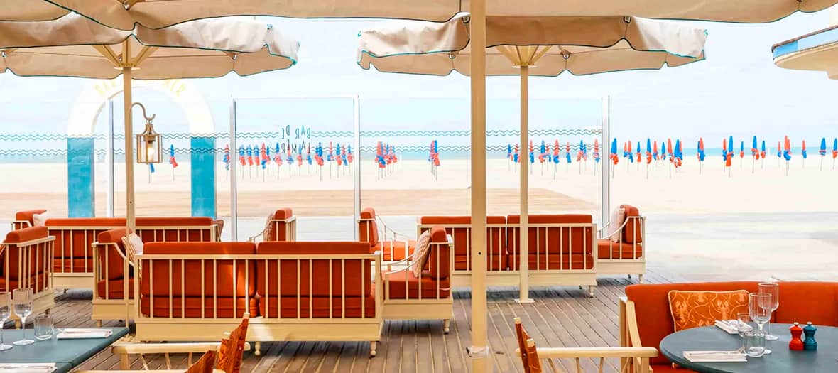 Terrasse face à la plage de Deauville avec vue sur les célèbres parasols rouges et bleus, symbole chic de la station balnéaire.